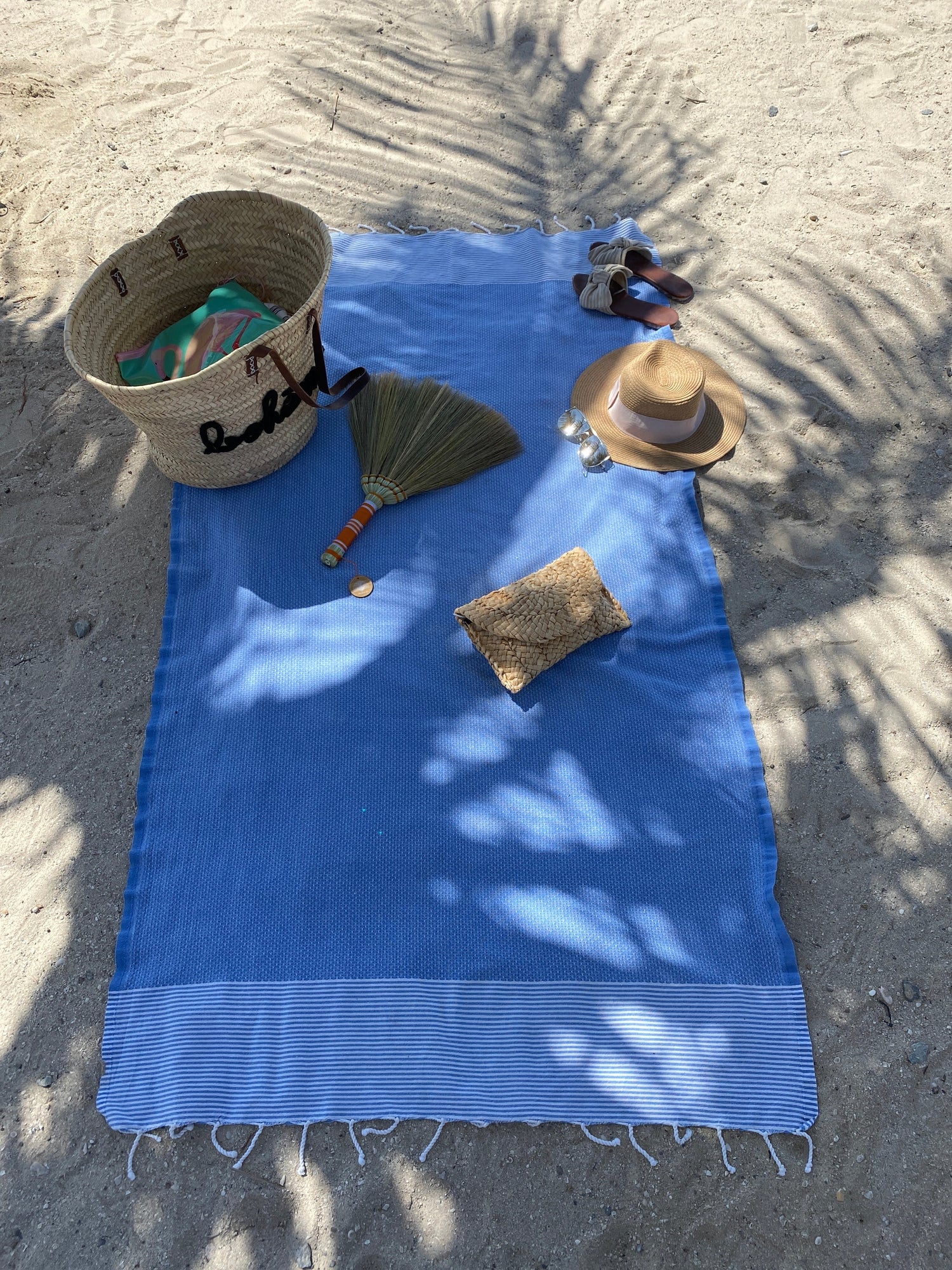 Blue towel on sand with beach accessories including a hat, bag, and fan.